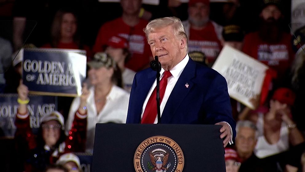 Politiker Donald Trump i kostym håller tal vid podium med presidentens sigill, publik i bakgrunden.