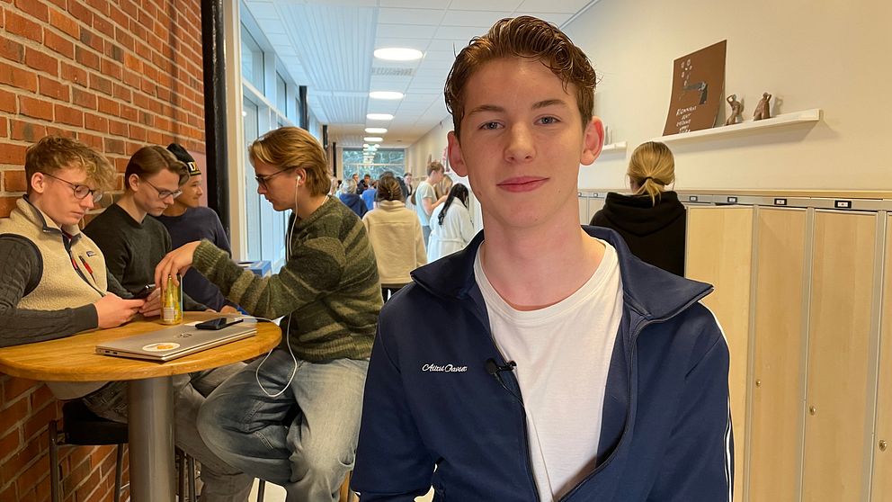 En skolkorridor med flera elever som sitter vid ett högt bord och samtalar medan en annan elev sitter närmare kameran. Korridoren har röda tegelväggar och skåp längs den motsatta väggen. Ljuset är starkt och naturligt från fönstren i bakgrunden.