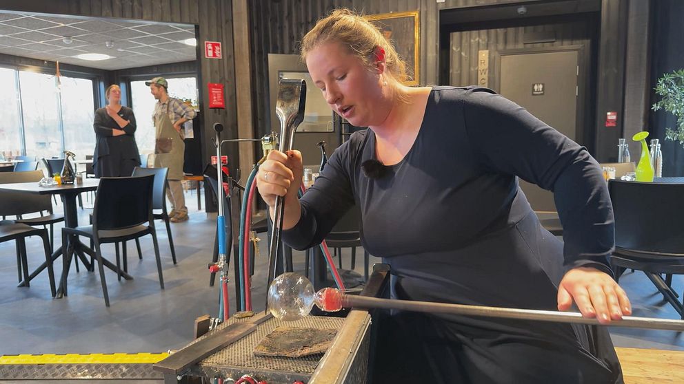 En person som arbetar med glasblåsning står vid en glasblåsarbänk. I förgrunden håller hon i ett glasverktyg och formar en glasboll. I bakgrunden syns två personer som tittar på, i en miljö med mörka väggar och moderna möbler.
