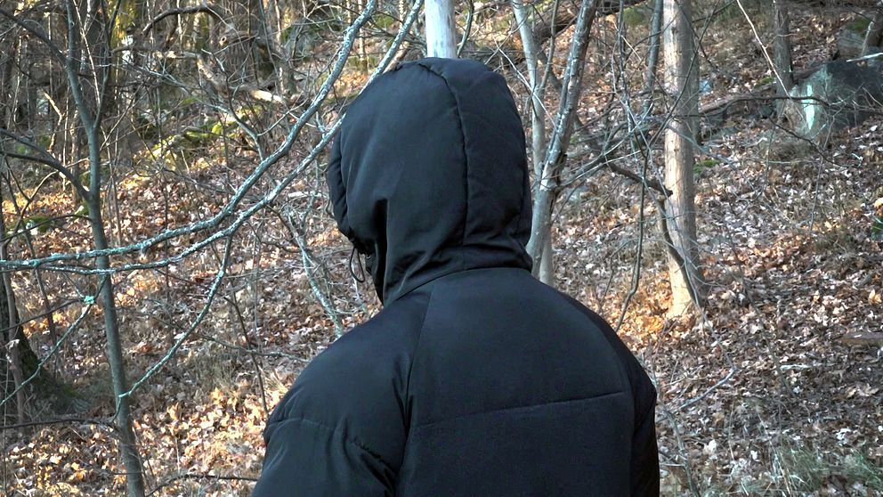En person med svart jacka och huva står med ryggen vänd mot kameran, omgiven av skog med löv och träd.