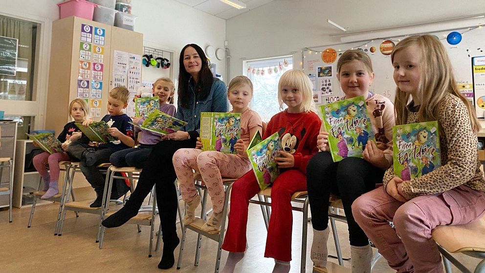 Barn sitter i en rad av stolar i en klassrumsmiljö, alla håller i böcker och ser engagerade ut. I mitten av raden sitter en vuxen. Rummet är dekorerat med färgglada affischer och material som tyder på en lärande miljö.