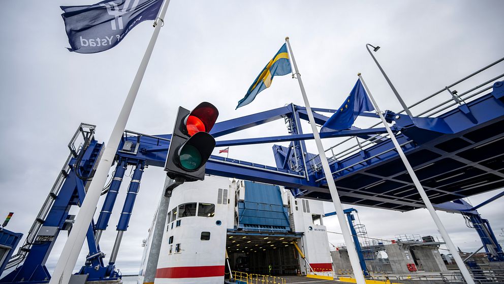 Färja som står vid en hamnterminal med nedfälld landgång för ombordstigning eller avstigning, omgiven av flaggor och ett trafikljus som visar rött ljus.