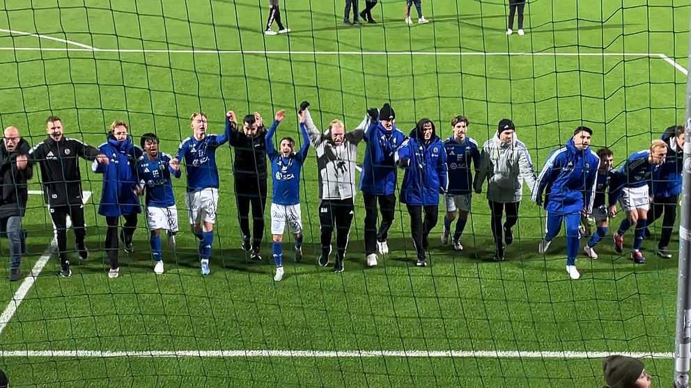Lagmedlemmar och tränare firar en seger på en fotbollsplan. De står i rad framför en nätad målbur, klädda i blåa och svarta träningskläder, med händerna lyfta i luften och leenden på läpparna. I bakgrunden syns andra personer som deltar i firandet. Gräset är grönt och planen ser välskött ut.