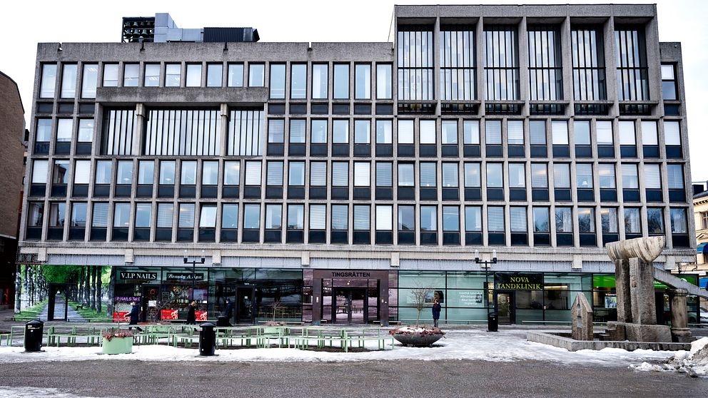 Modern byggnad med stora fönster och en symmetrisk fasad, belägen på en gatumiljö med butiker i bottenvåningen. I förgrunden finns en fontän och sittplatser, omgiven av snö.