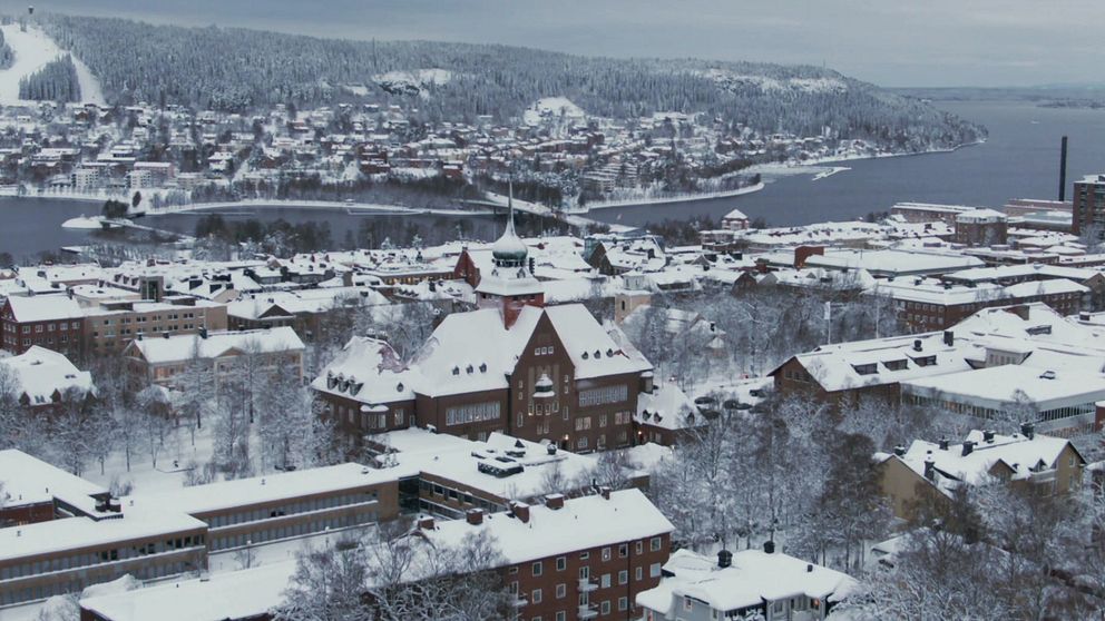 En panoramabild över en snötäckt stad med traditionell arkitektur, belägen vid en sjö. I förgrunden syns flera byggnader med snöbelagda tak, inklusive en stor, historisk byggnad med en tornspira. I bakgrunden reser sig en kuperad, snötäckt landskap med fler hus och träd, och sjön breder ut sig till höger.