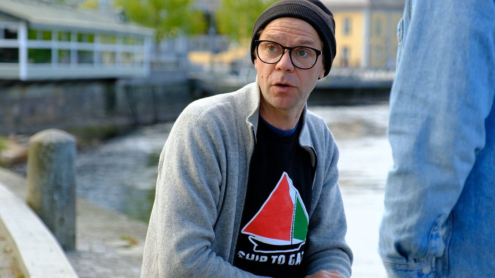 Person med grå kofta och svart t-shirt sitter utomhus vid vattnet, samtalar med annan person.