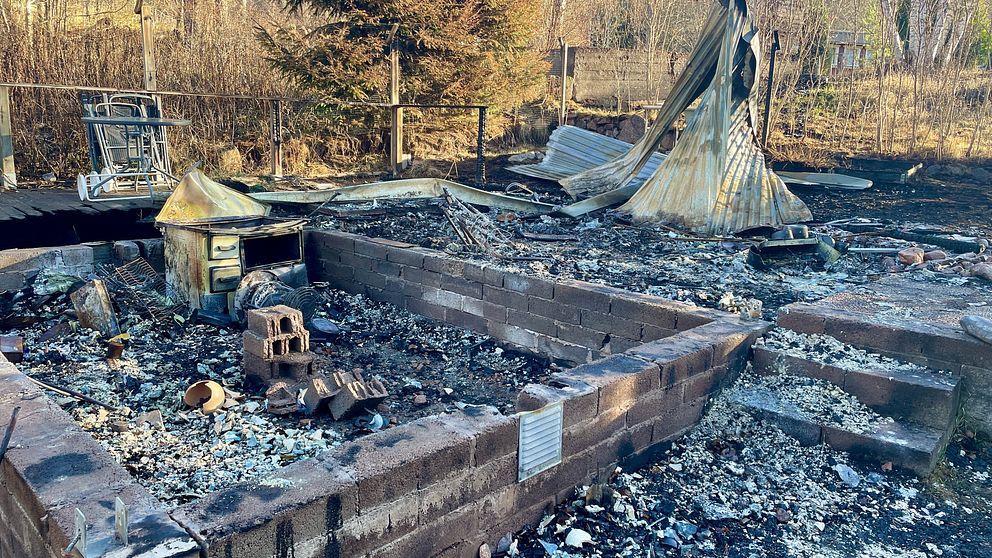 Rester av en utbränd byggnad på en tomt, med förkolnade tegelväggar och förvridna metallstrukturer. Bakgrunden visar träd och en åker.