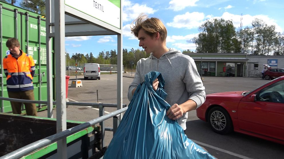 Person lämnar avfallspåse på återvinningsstation, personal med reflexväst i bakgrunden.