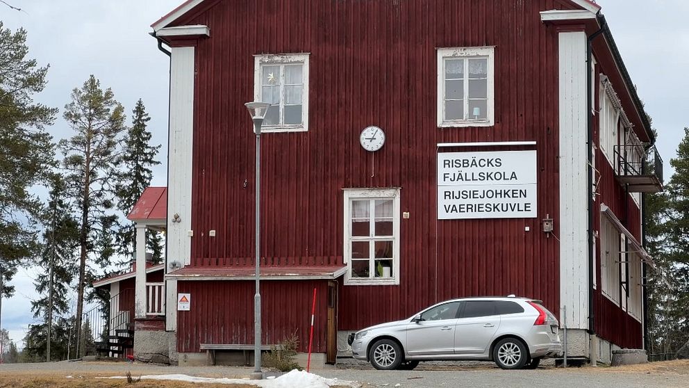 Röd byggnad, Risbäck fjällskola. Silverfärgad bil parkerad. Träd i bakgrunden.