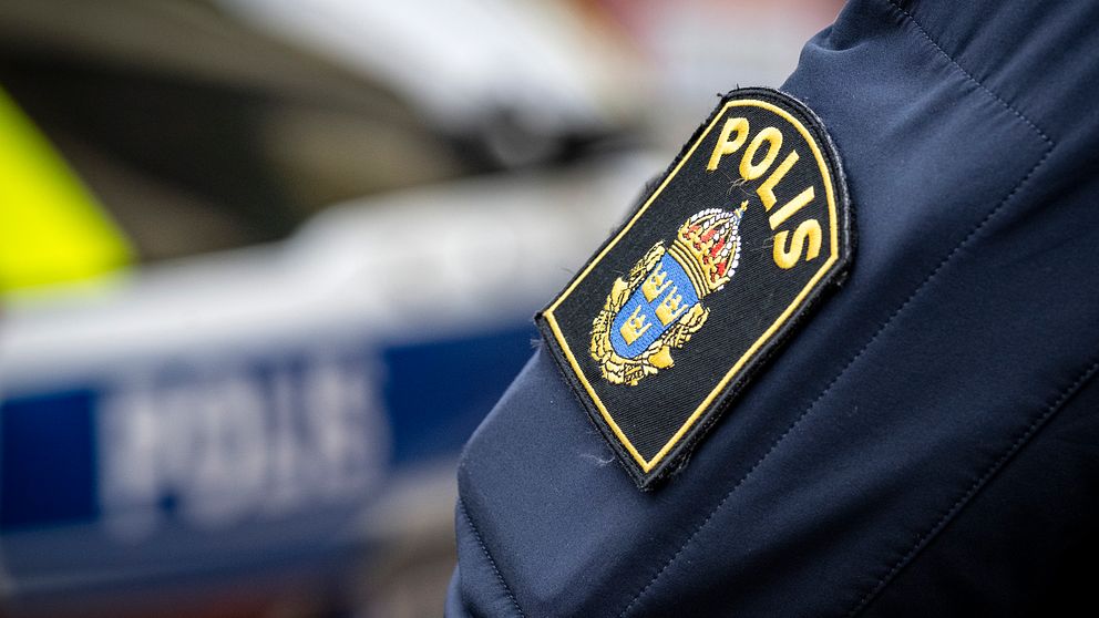 En närbild av en polisuniform där ett tydligt insynskt patch med texten ”POLIS” är synligt på ärmen. I bakgrunden syns en polisbil.