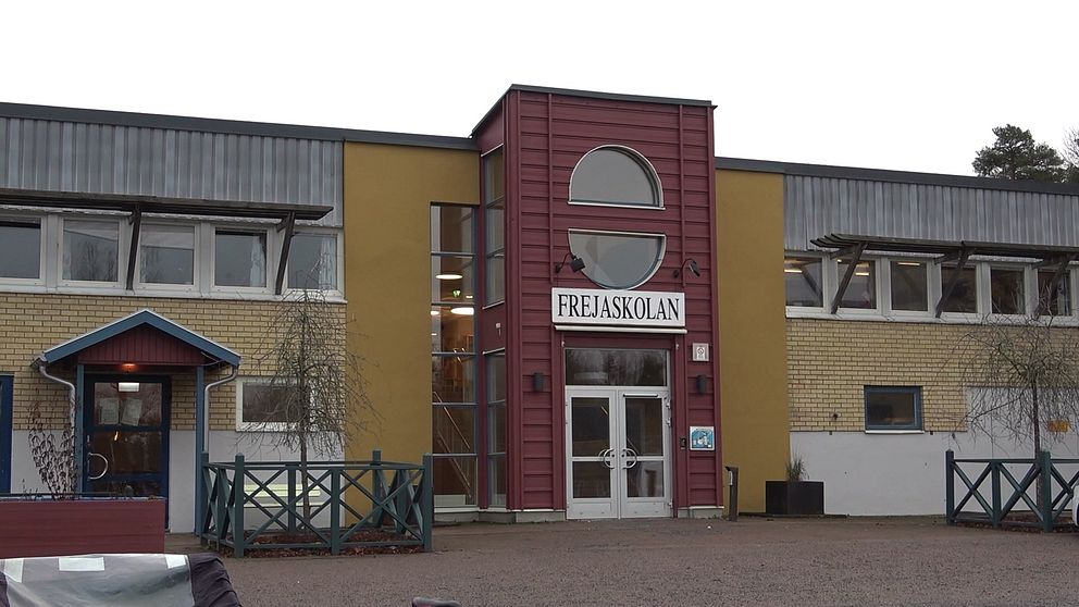 Fasaden av Frejaskolan, en modern byggnad med en röd och beige exteriör. Ingången är centralt placerad och omgiven av stora fönster. Omgivningen är lugn, med en grönskande gård och en trägrind synlig till vänster.