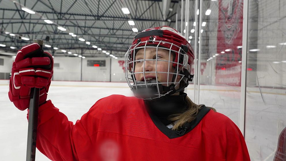 Ishockeyspelare i röd tröja och hjälm står vid sargen på en ishall.