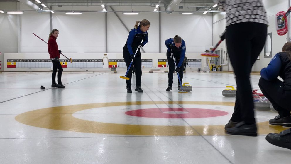 Personer spelar curling på en inomhusbana. Sopare vid sidan om stenen.