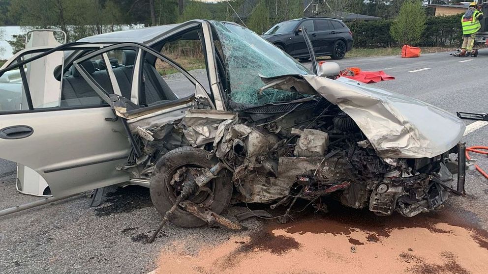 Skadad bil efter en olycka, med kraftigt krockad front och bakhjul synliga. Området runt bilen är avspärrat med säkerhetsvästar synliga och sand spridd på vägbanan för att absorbera vätskor. I bakgrunden syns en sjö och träd.