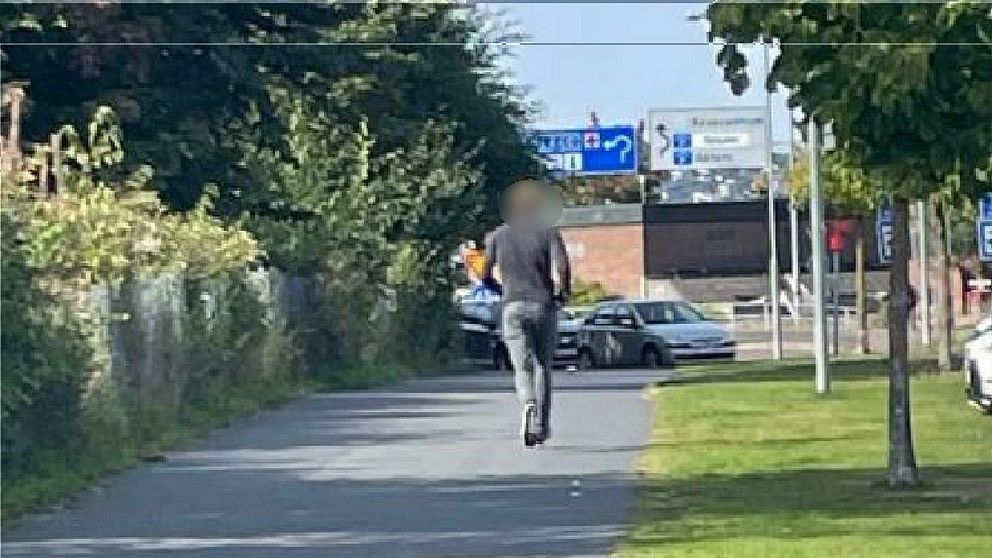Person går längs trottoar kantad av bilar och träd i solljust område.