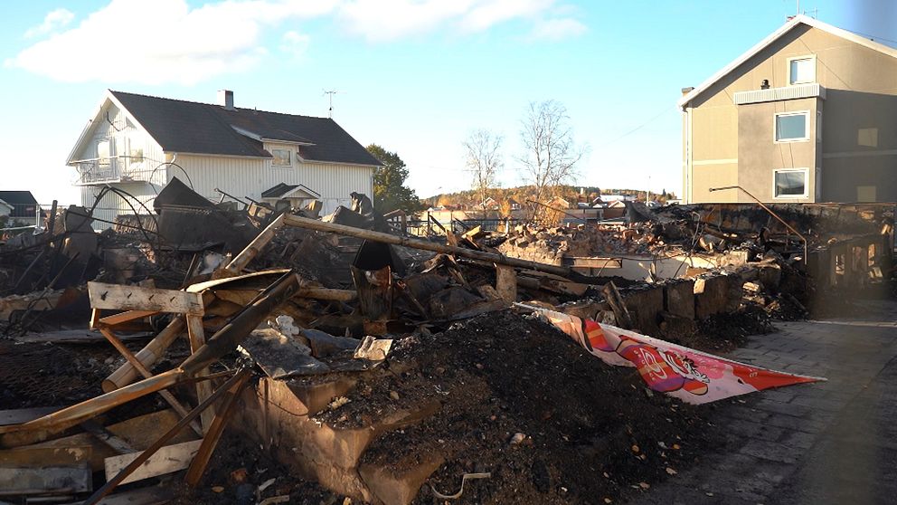 Detaljerad vy av en plats som drabbats av en brand, med jämna ruiner och nedbrunna byggnadsdelar. I bakgrunden syns en intakt byggnad och ett ljust blått himmel. Rester av bråte och aska är spridda över området, vilket vittnar om den förödelse som orsakats. En del av en färgglad skylt kan skymtas bland rubbningen.