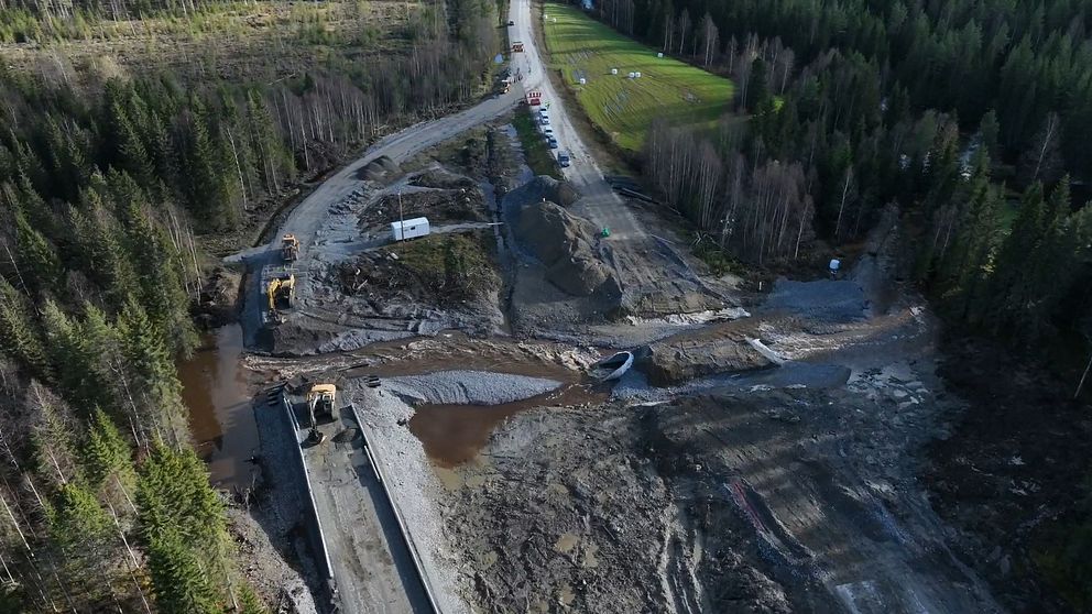 Vy över en byggarbetsplats med grönskande skog runt omkring. I bilden syns nya vägar som runnit sönder och maskiner som arbetar. En del av området är blöta och leriga, och det finns en sektion där vattnet rinner.