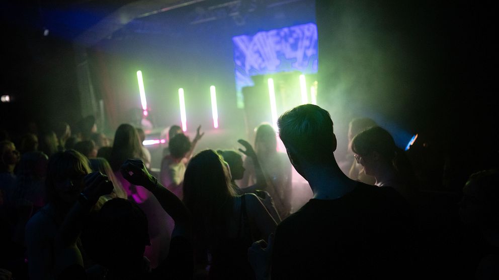 Nattklubbs scen med festande människor under neonljus och dimma. Flera personer dansar framåt mot scenen där färgglada ljus blinkar. Atmosfären är livlig och energisk.