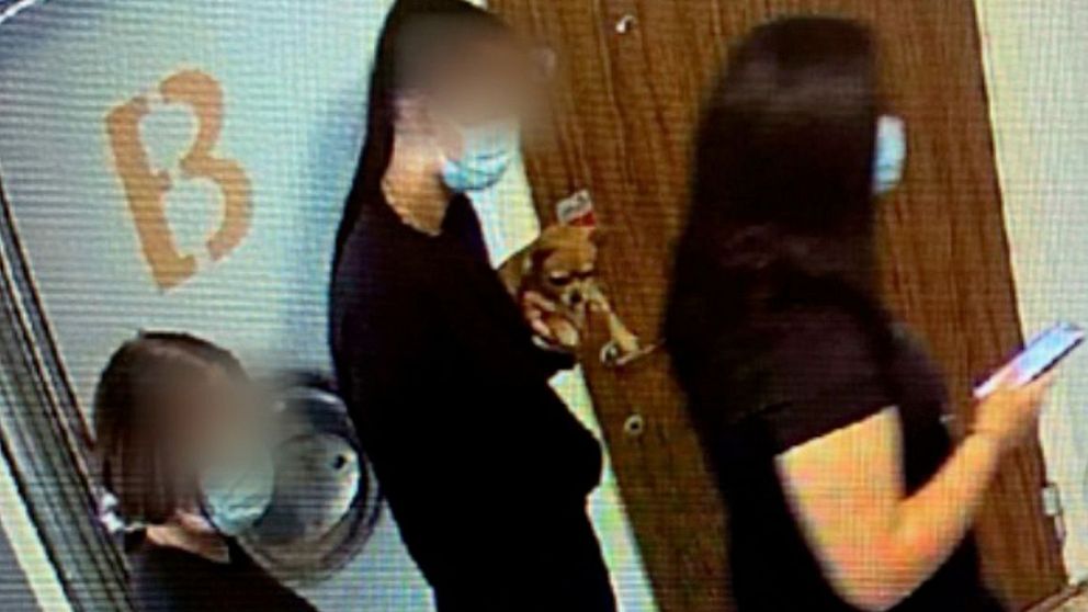 Tre personer, med ansiktena suddiga för anonymitet, står i en rad i en korridor utanför en dörr. De bär ansiktsmasker och kläder i mörka nyanser. Personen längst fram håller en mobiltelefon. En stor bokstav ”B” syns på väggen.