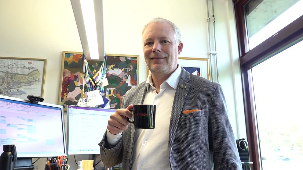 Politiker står vid ett skrivbord och håller en svart kopp med Lejonens logga på. I bakgrunden syns två datorskärmar som visar arbetsrelaterad information, tillsammans med konstverk på väggen. Rummet har stora fönster som släpper in naturligt ljus.