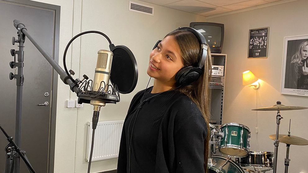 En person står framför en mikrofon i en inspelningsstudio, iklädd hörlurar. I bakgrunden syns ett trumset samt affischer på väggarna. Rummet har en professionell atmosfär med dämpad belysning.