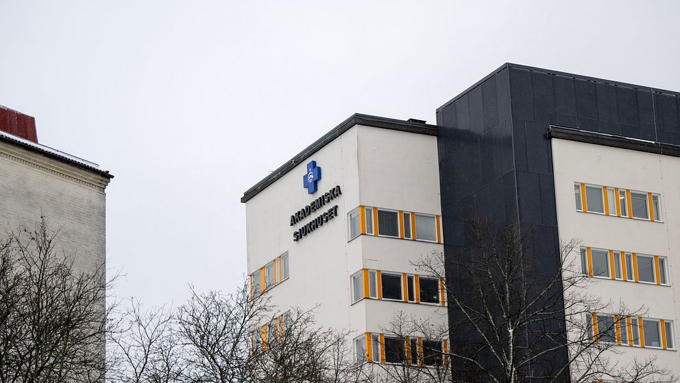 Sjukhusbyggnad på Akademiska med logotyp och gula fönster, trädgrenar i förgrunden.