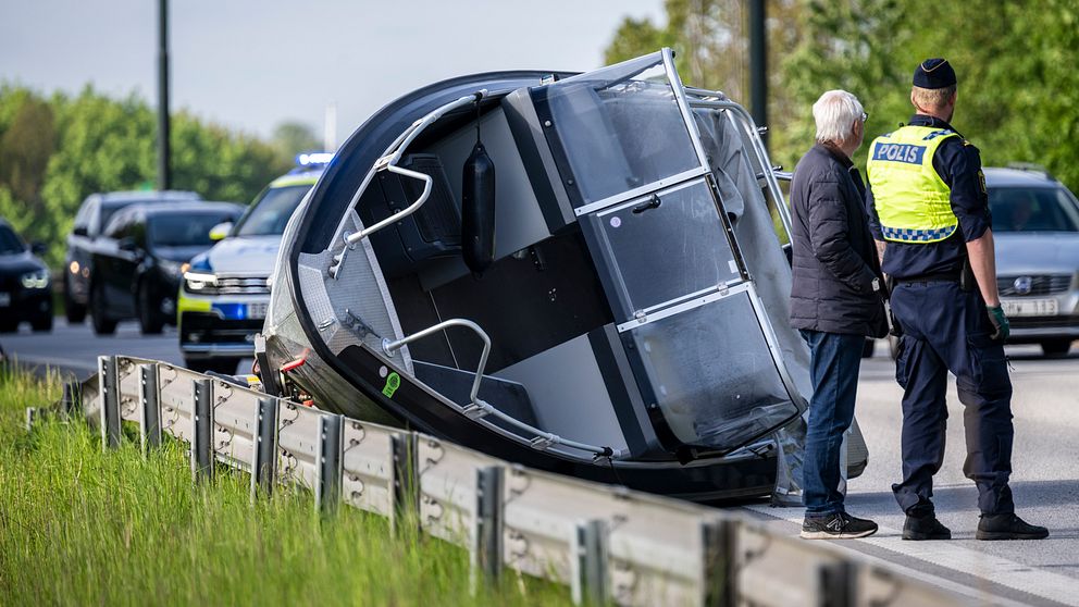 Bil har vält vid vägkant, polis och civilperson står bredvid, kö med bilar i bakgrunden.