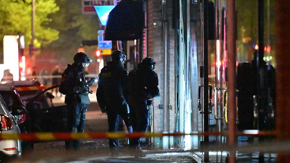Tre poliser i mörka kläder står och ser på en skadad glasdörr.
