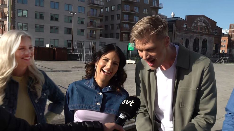 Reporter håller ut en mikrofon från SVT till tre personer på ett torg med byggnader i bakgrunden.