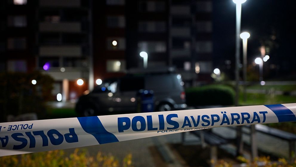 Polisavspärrning med texten ”POLIS AVSPÄRRAT” i förgrunden, med en bil och flerfamiljshus i bakgrunden, belyst av gatulampor under natten.