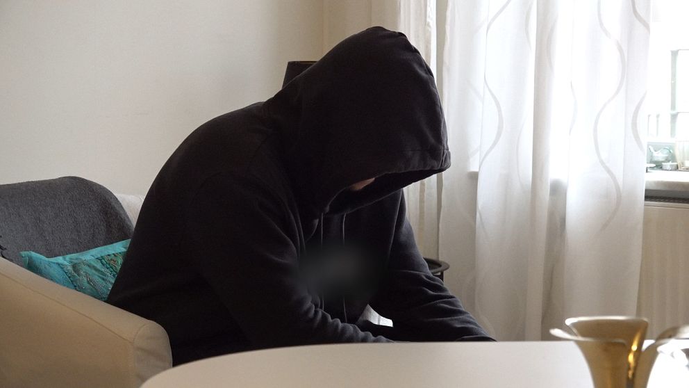 En person med huvtröja sitter på en soffa, lutad framåt med huvudet sänkt. Rummet har vitt ljus som strömmar in genom ett fönster, och en ljusstake syns på bordet framför. Det finns också en färgad kudde på soffan. Atmosfären verkar lugn och stillsam.