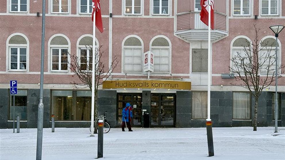 Entrén till Hudiksvalls kommunhus, där snö ligger på marken och två personer syns utanför. Klassiska fönster i grått och rosa syns på byggnadens fasad, med svenska flaggor som svajar.