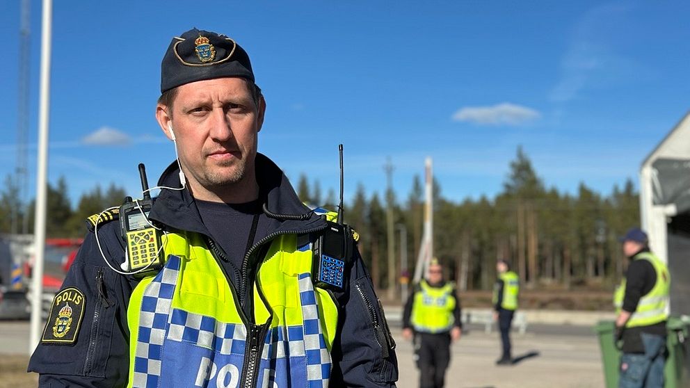 Polis med insatsväst står utomhus vid ett skogsområde, andra poliser i bakgrunden.