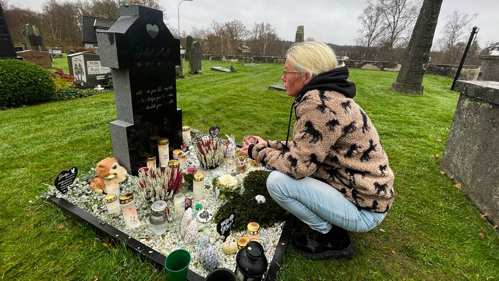 Camilla Hall knäböjer vid sin sons gravsten på en kyrkogård i Örkelljunga, omgiven av blommor, ljus, och minnessaker. Gräset är grönt och omgivningen är lugn, med träd och andra gravar i bakgrunden. Det är en molnig dag.