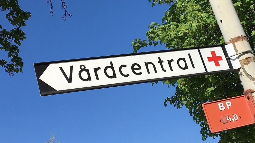 Vägskylt som det står ”Vårdcentral” på med en klarblå himmel i bakgrunden.