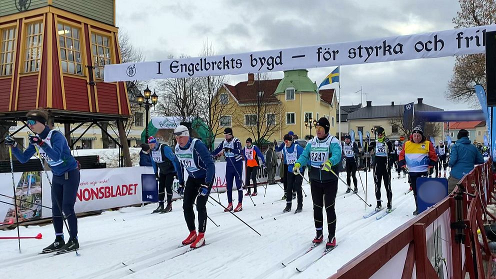 Skidåkare deltar i ett långlopp i en svensk stad, med en banderoll ovanför spåret. Flera åkare i nummerlappar syns i förgrunden, medan åskådare och färgglada byggnader finns i bakgrunden. En svensk flagga vajar bland träden.