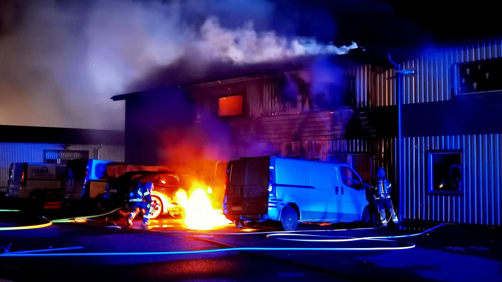 På bilden syns en brinnande bil. Det är mörkt ute. Bilen står utanför en industrilokal. En brandman är framme vid elden och försöker släcka den.