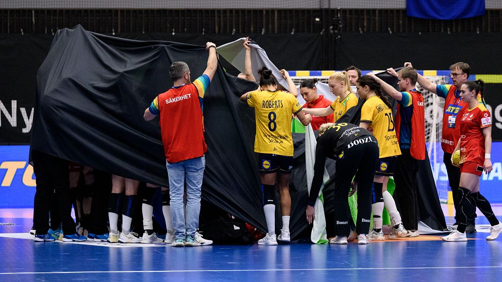 Handbollsspelare omringade av en svart duk på en inomhusplan.