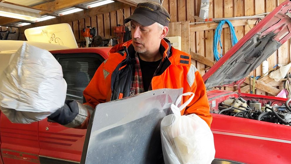 Person i orange jacka håller plastpåsar och bildel framför röd bil med öppen motorhuv.