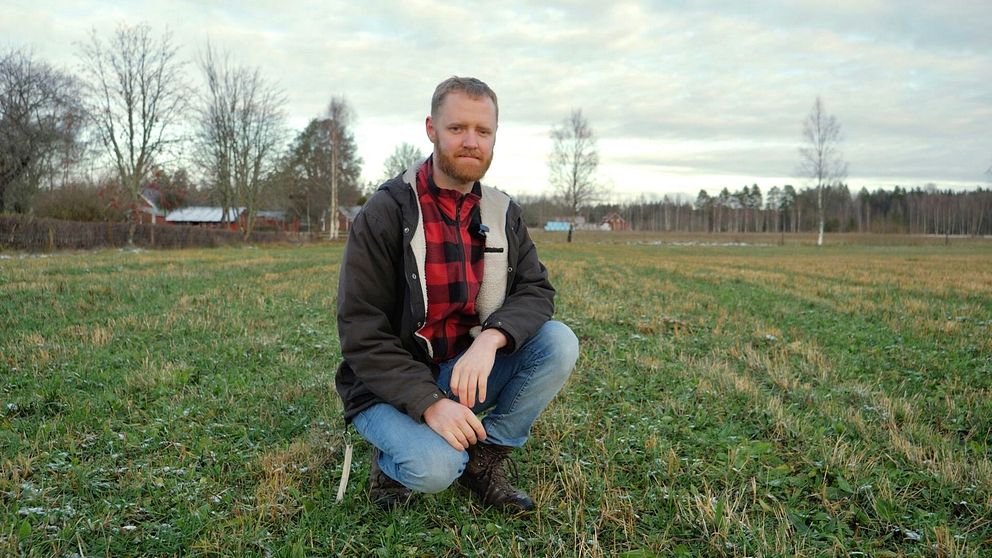 En person sitter på knä i ett öppet fält med grön gräs och några spridda träd i bakgrunden. Himlen är delvis molnig och ger en stillsam atmosfär. I bakgrunden syns några hus på avstånd.