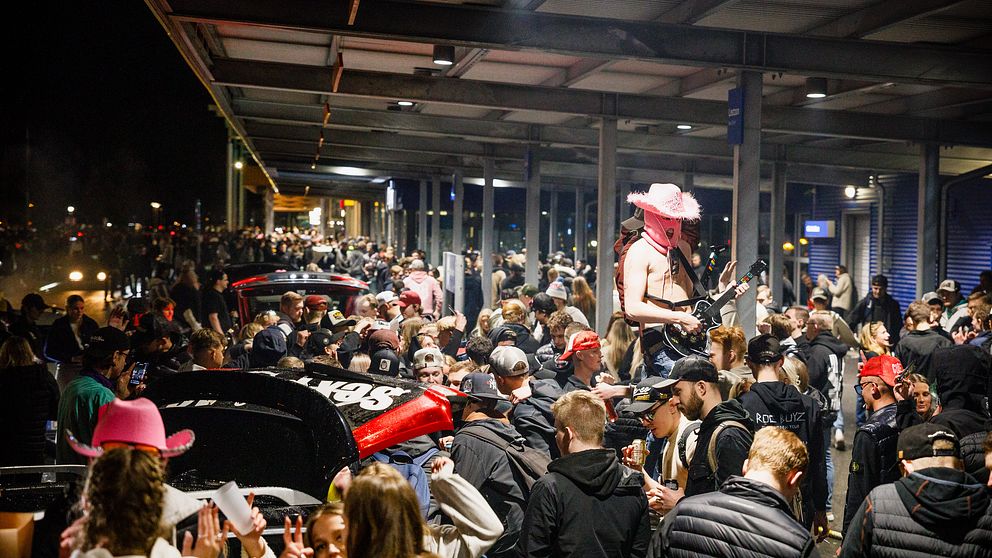 Folkmassa vid A6-parkeringen i Jönköping, en kille spelar gitarr iklädd ansiktsmask.