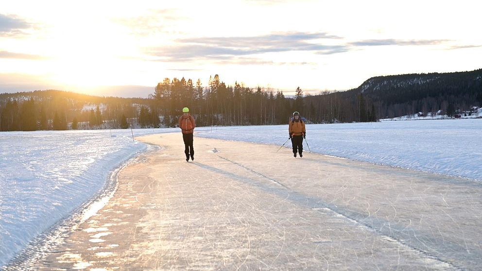 Två personer åker skridskor på en frusen sjö med solnedgång i bakgrunden. Den vänstra personen bär en gul hjälm och orange jacka, medan den högra personen har en orange jacka och stavar. Området är omgiven av snö och träd, vilket ger en vinterlig atmosfär.