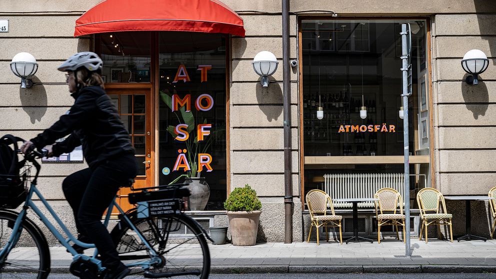 En cyklist passerar en restaurang med namnet ”ATMOSFÄR” som har en röd markis och utomhusmöbler. Fönstren har dekorativa ljus och en grön växt är placerad vid entrén.