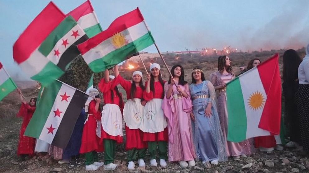Grupp personer i festkläder håller kurdiska och syriska flaggan.