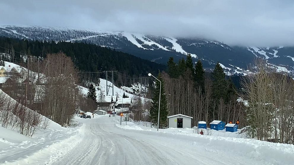 Snöig landsväg leder mot skidbackar och berg täckta av snö under molnig himmel.