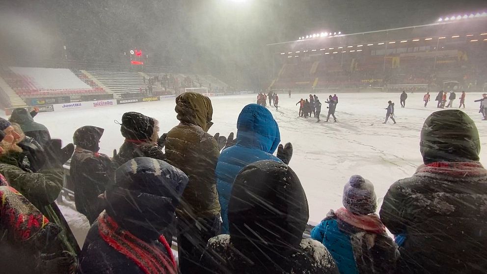 En fotbollsmatch pågår i kraftigt snöfall, där åskådarna i förgrunden är insnurrade i varma kläder och håller sig i skydd från vädret. I bakgrunden visas spelare som rör sig över planen, omgiven av snö. Ståplatsläktaren är delvis synlig, och belysningen från arenan kämpar för att tränga igenom snöfallet.