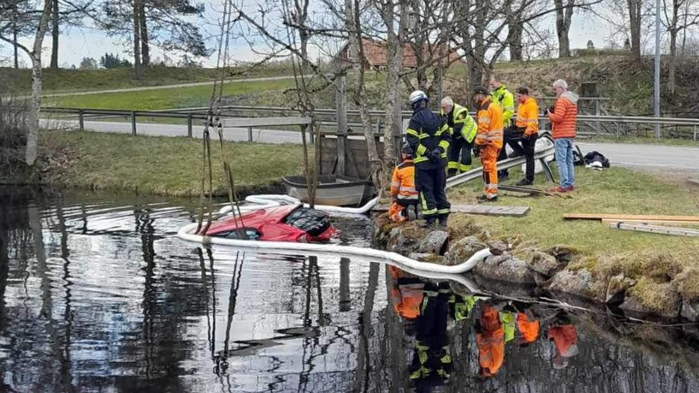 En röd bil ligger i vattnet, bara bakdelen och taket syns. Till vänster syns en lastbil som försöker bärga bilen. Mellan bärgaren och bilen står flera personer.