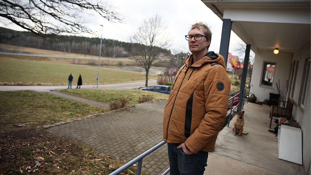 En man står på en veranda med utsikt över ett landskap med en asfalterad väg och åkrar. En hund sitter bredvid honom vid husets yttervägg. I bakgrunden promenerar två personer längs gångvägen.
