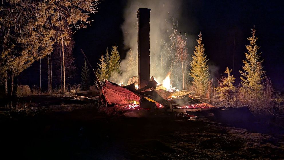 Brinnande trähus på natten, omgiven av träd och rök. Bara murstocken står kvar.