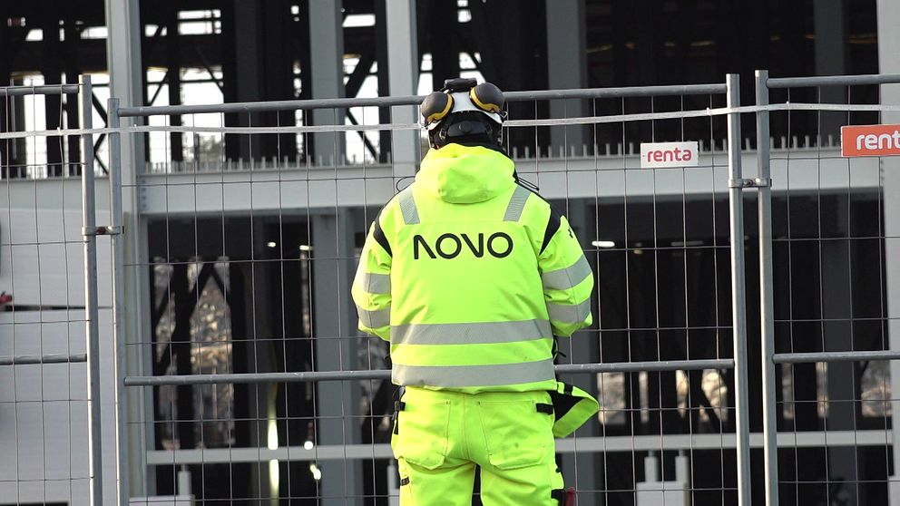 Byggarbetare i reflexväst står vid en avspärrning framför en byggnad under konstruktion vid bygget av Volvo Cars satsning på batterifabrik i Göteborg.